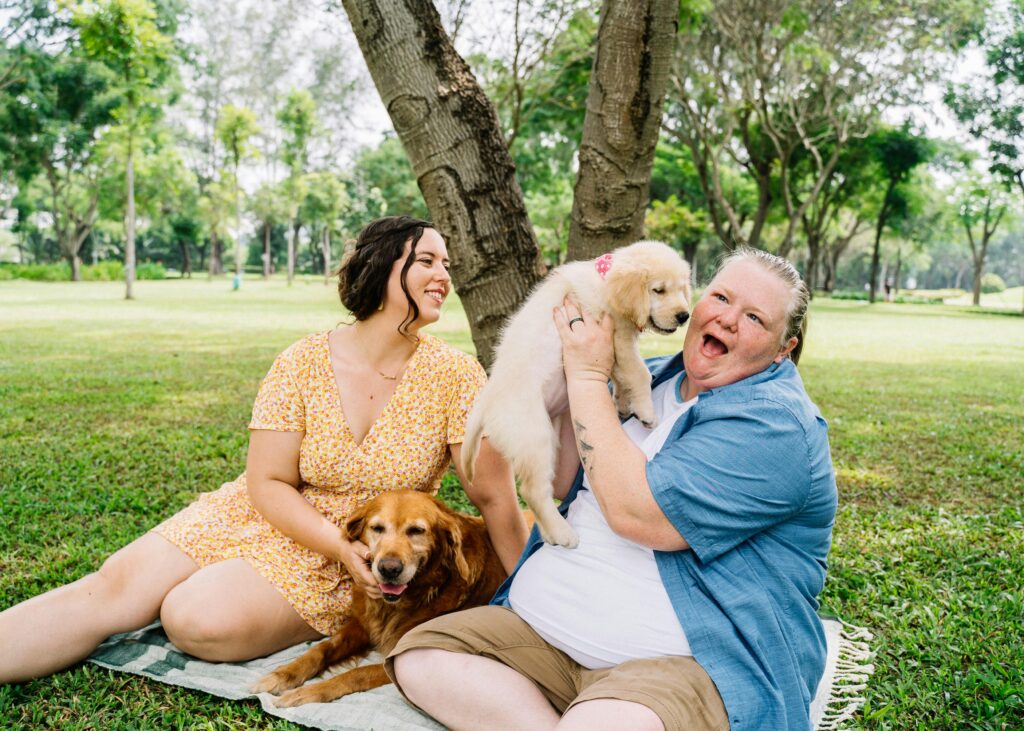 A joyful couple sitting with their Golden Retriever and a puppy in a lush green park.