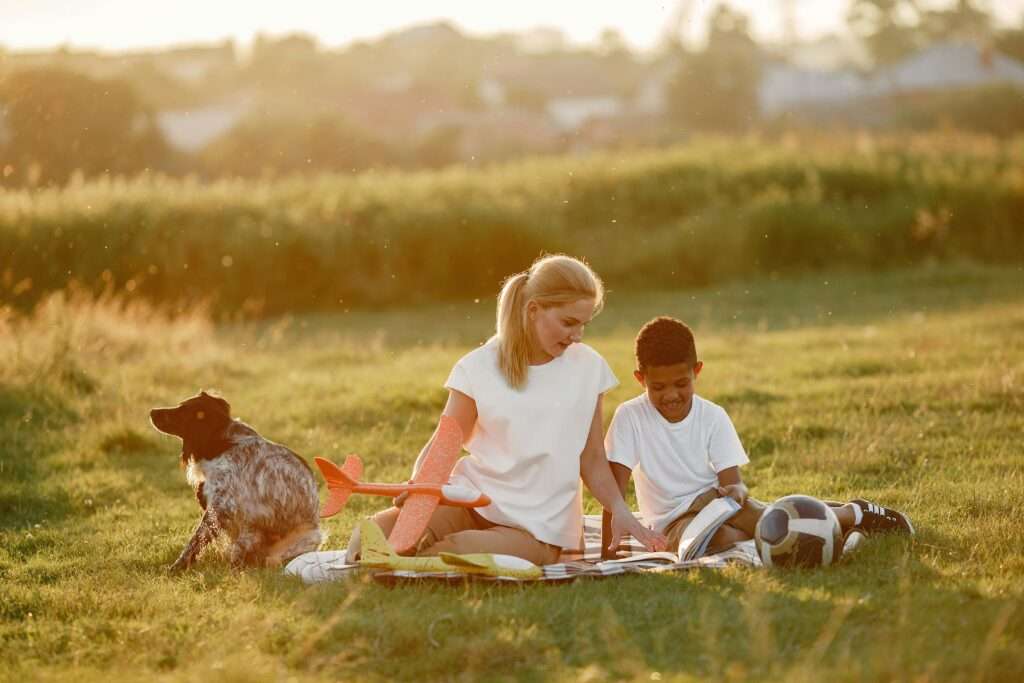 A woman and young boy relax with a dog and toys in a sunny meadow.
